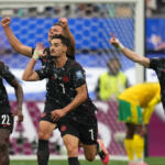 Soccer players in black uniforms celebrate a goal on the field, center player #1 signaling with a raised hand while teammates run with raised arms near the goal.