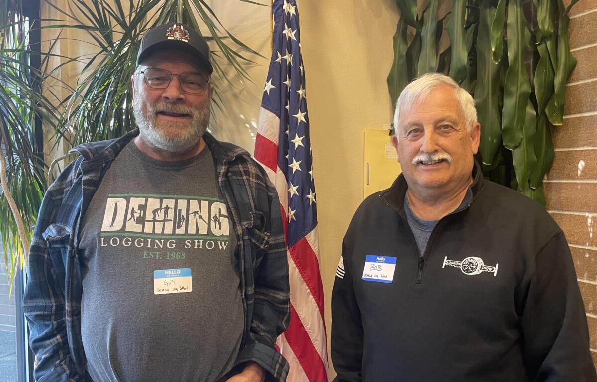 Two men standing indoors with an American flag in the background, both wearing name tags and casual clothing.