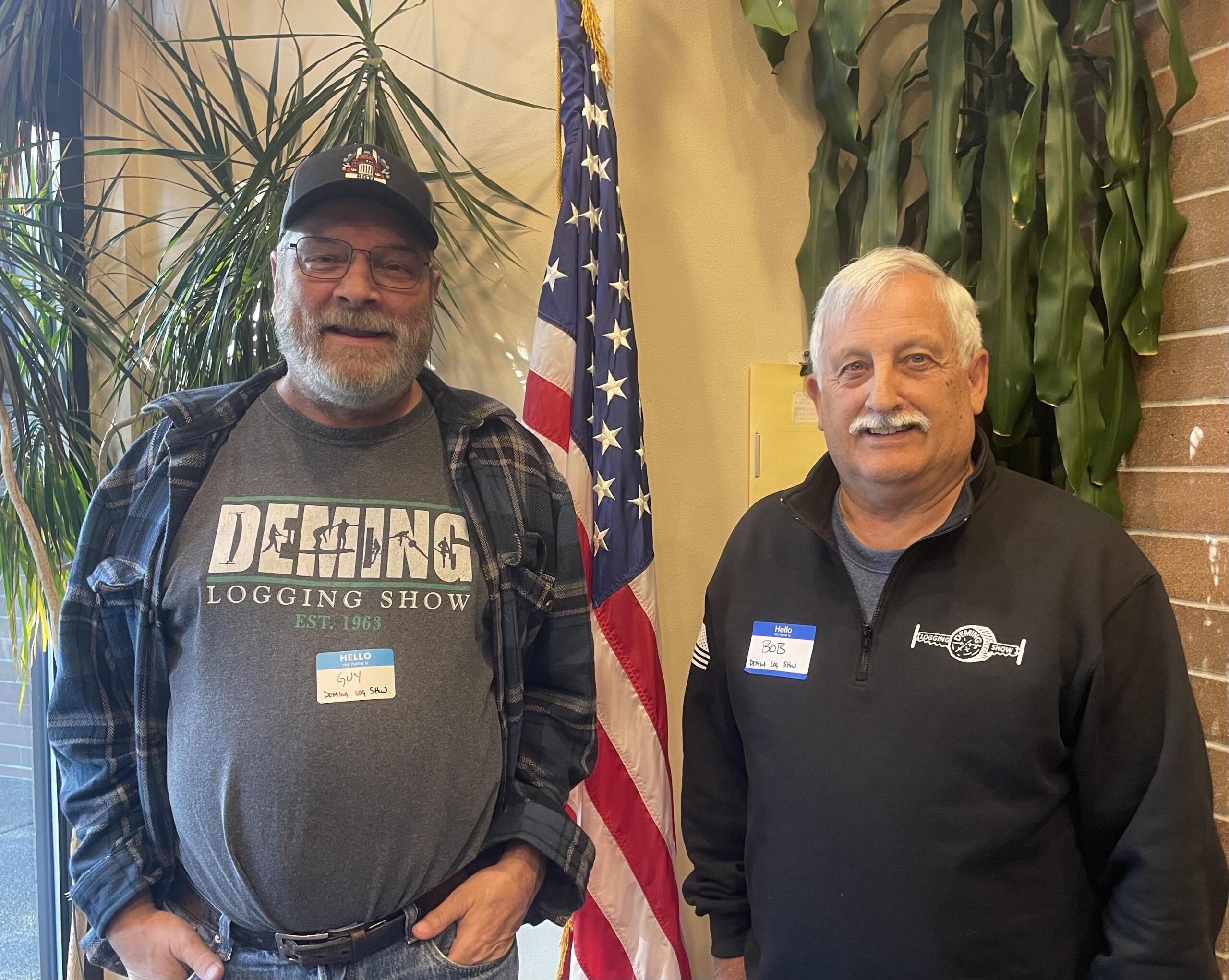 Two men standing indoors with an American flag in the background, both wearing name tags and casual clothing.