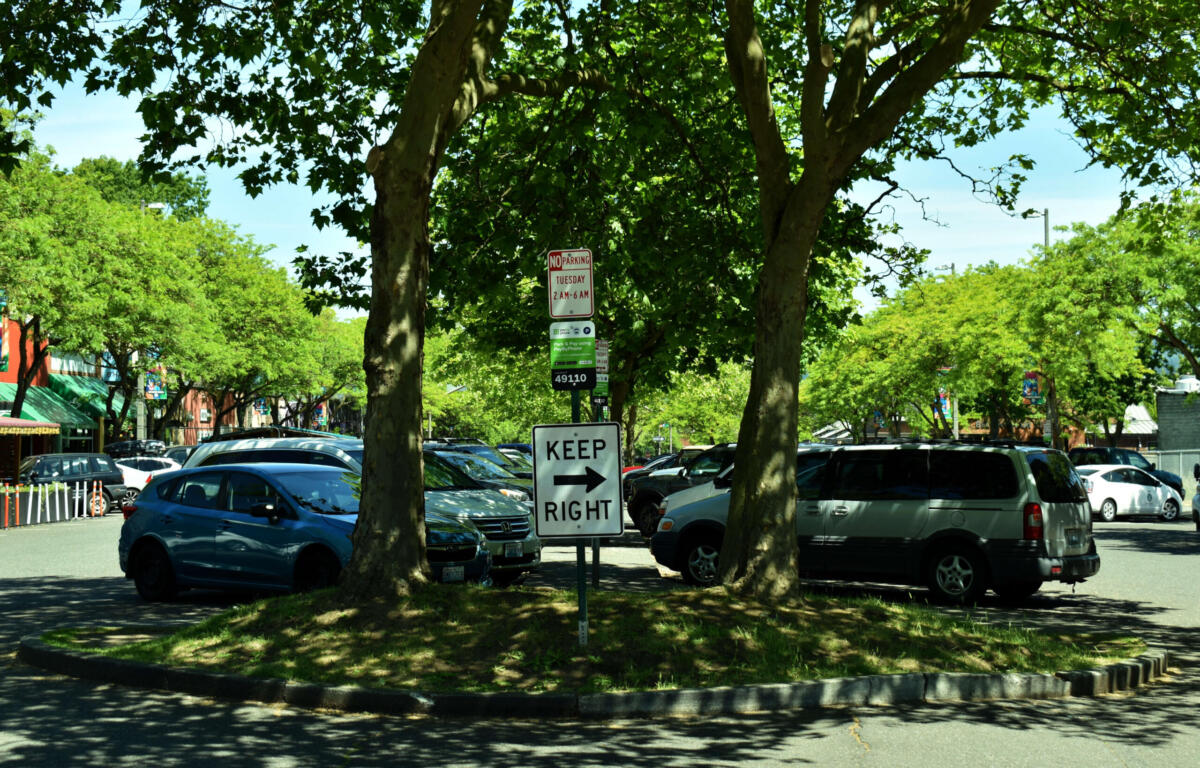 Parking lot under leafy trees with a prominent 'KEEP RIGHT' sign in the foreground and parked cars along the curb beneath a sunny sky.