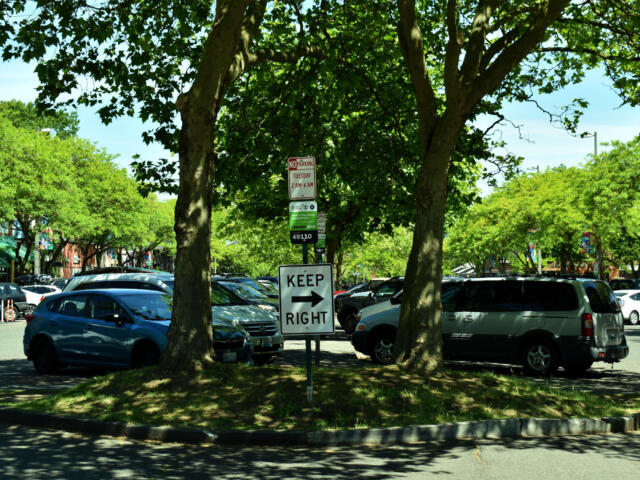 Parking lot under leafy trees with a prominent 'KEEP RIGHT' sign in the foreground and parked cars along the curb beneath a sunny sky.