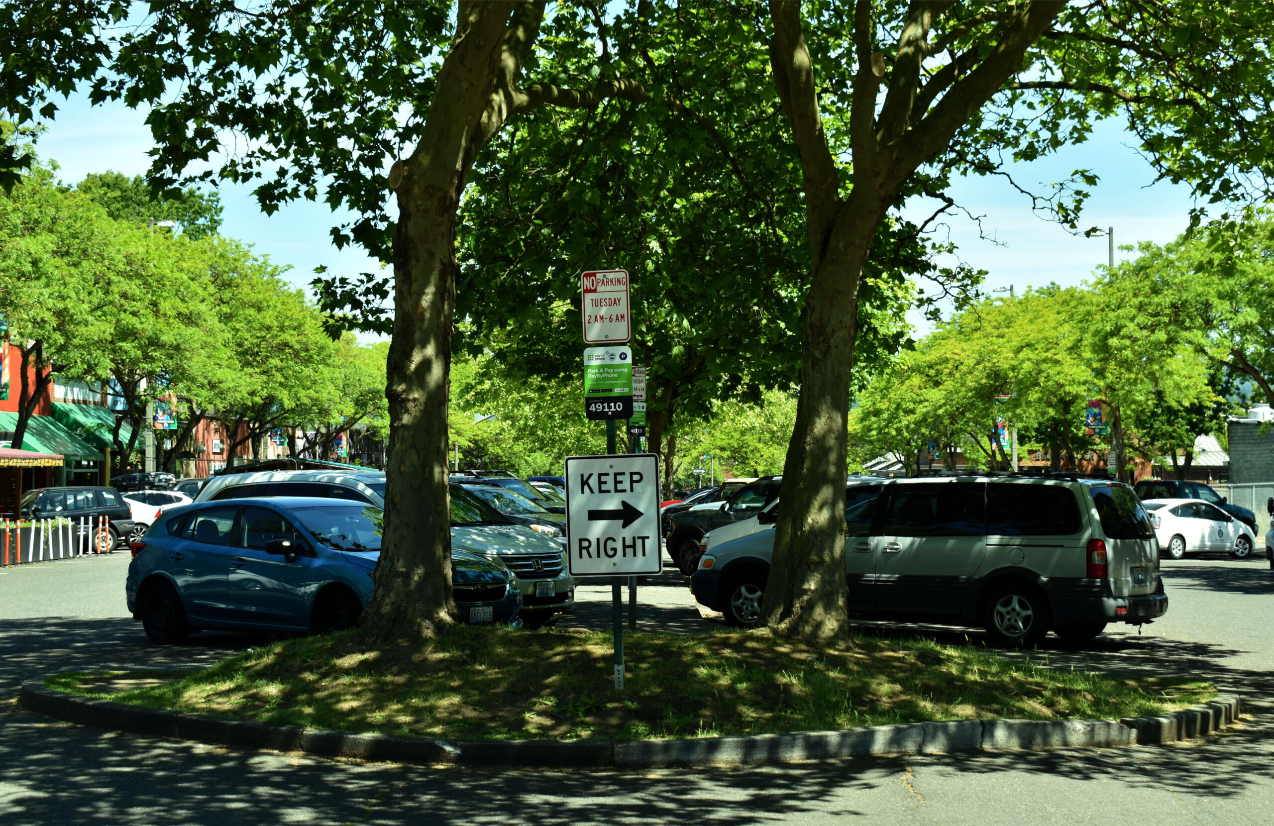 Parking lot under leafy trees with a prominent 'KEEP RIGHT' sign in the foreground and parked cars along the curb beneath a sunny sky.