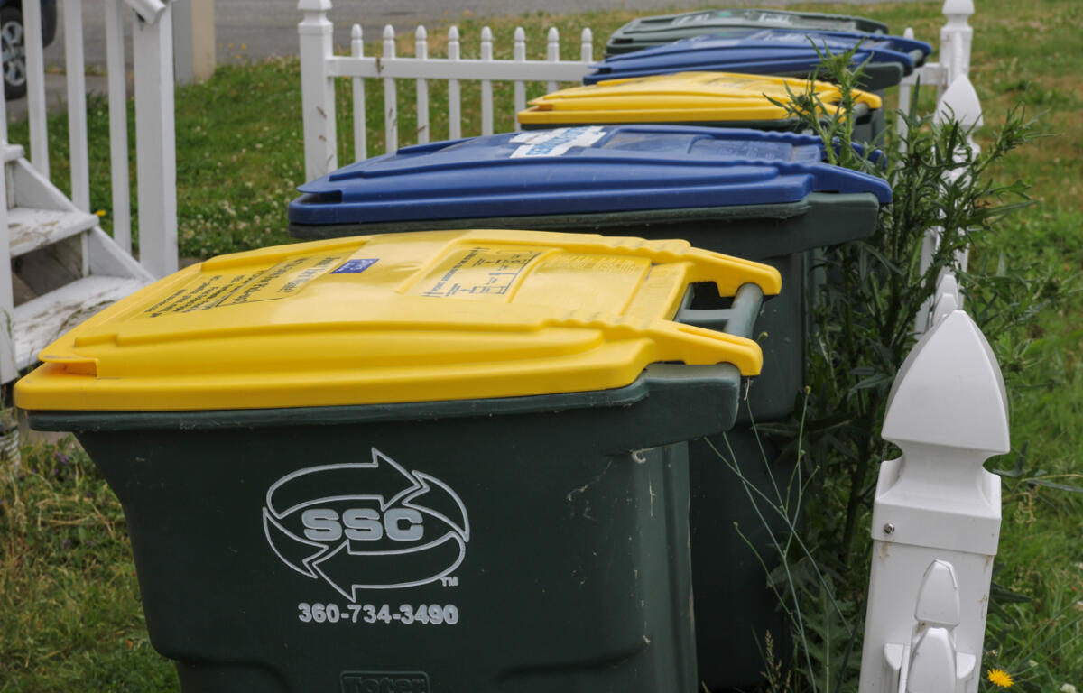 Row of residential waste and recycling bins with yellow, blue, and green lids along a white picket fence.