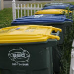 Row of residential waste and recycling bins with yellow, blue, and green lids along a white picket fence.
