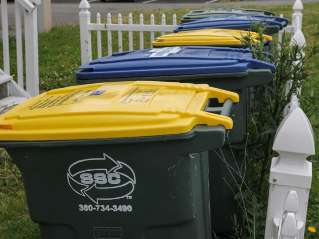 Row of residential waste and recycling bins with yellow, blue, and green lids along a white picket fence.