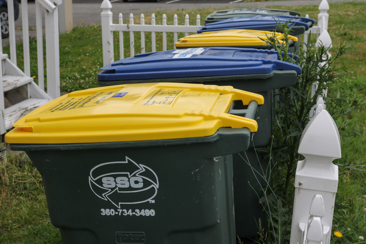 Row of residential waste and recycling bins with yellow, blue, and green lids along a white picket fence.