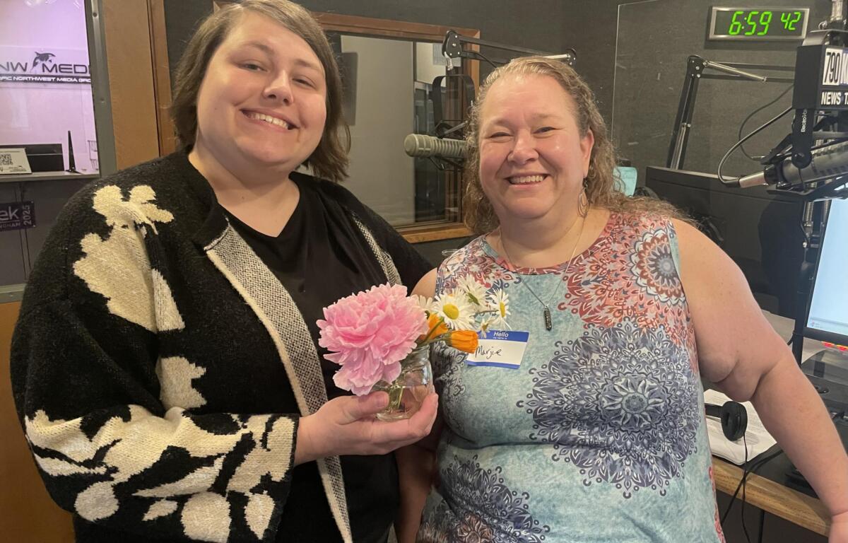 Two women smiling and posing in a radio studio, one holding a bouquet of pink flowers in front of them. Mic and equipment visible in the background.