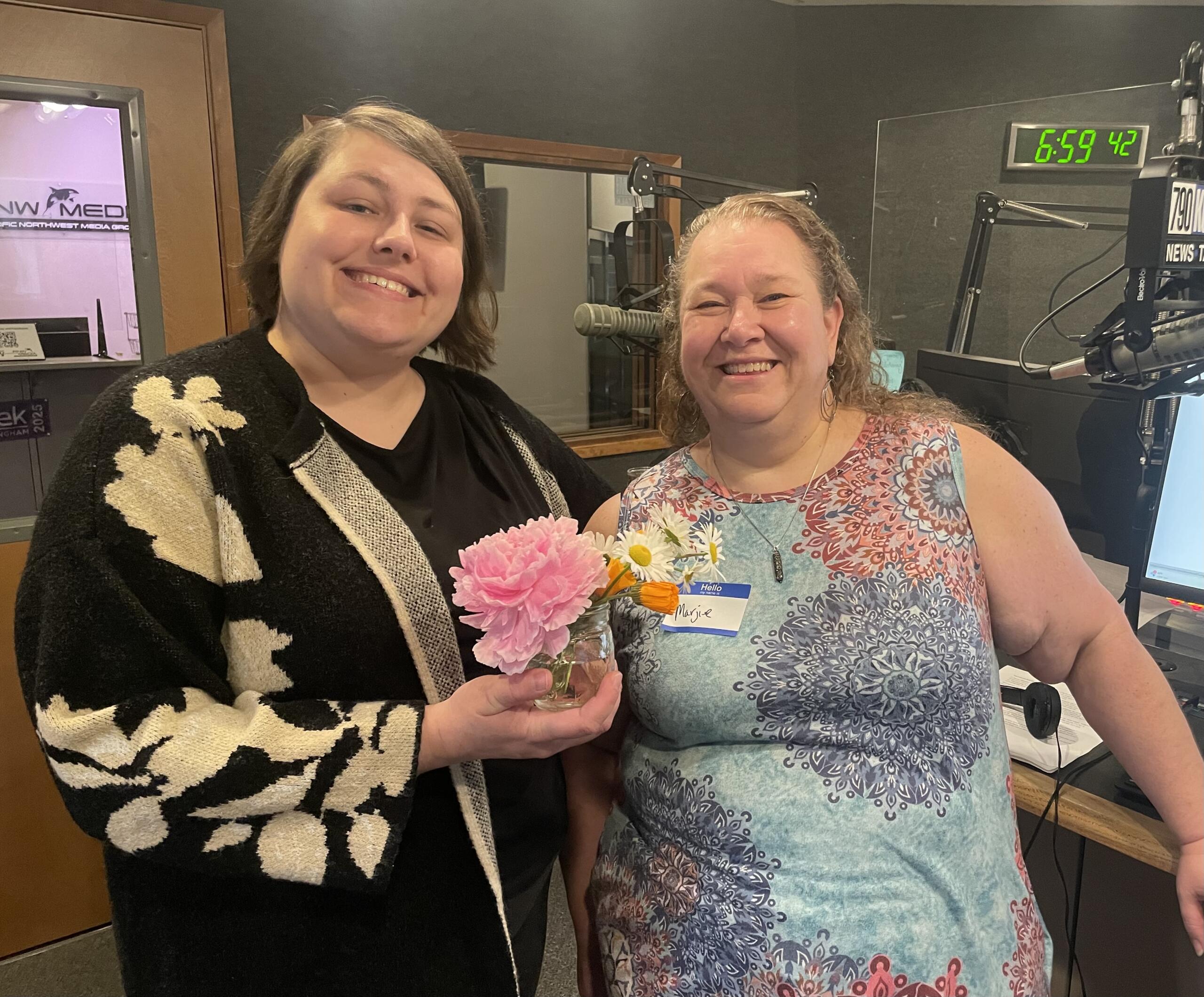 Two women smiling and posing in a radio studio, one holding a bouquet of pink flowers in front of them. Mic and equipment visible in the background.