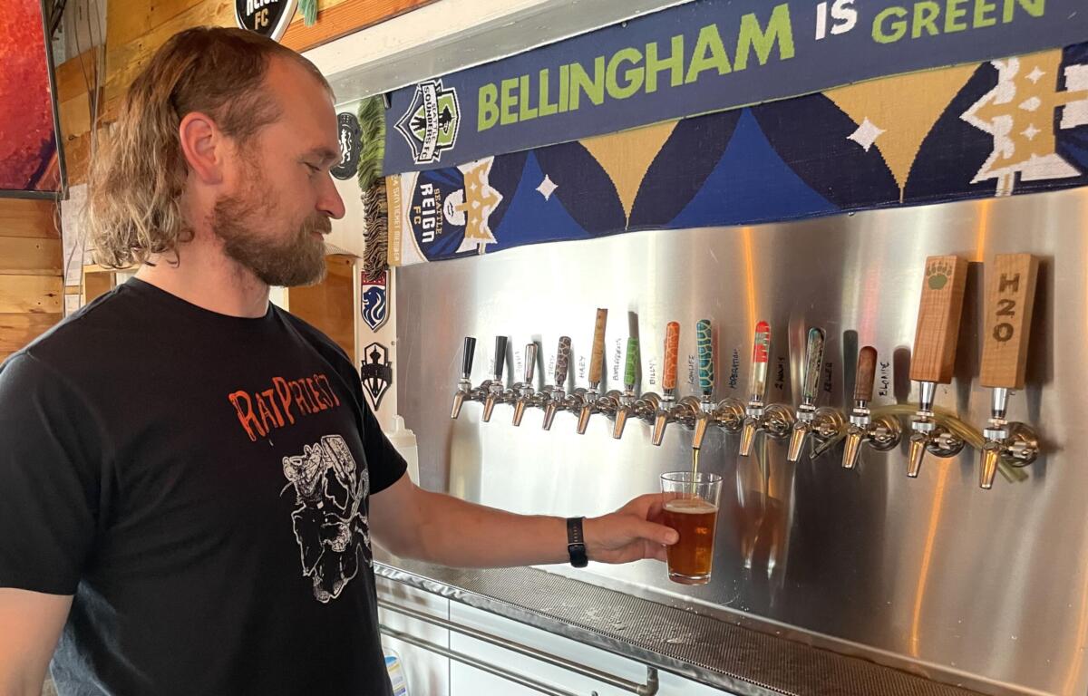 Man with long hair and beard in a black t‑shirt pouring beer into a glass at a row of taps in a bar, with a blue banner reading 'BELLINGHAM IS GREEN' in the background