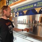 Man with long hair and beard in a black t‑shirt pouring beer into a glass at a row of taps in a bar, with a blue banner reading 'BELLINGHAM IS GREEN' in the background