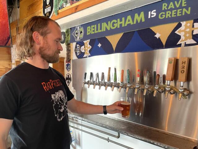 Man with long hair and beard in a black t‑shirt pouring beer into a glass at a row of taps in a bar, with a blue banner reading 'BELLINGHAM IS GREEN' in the background