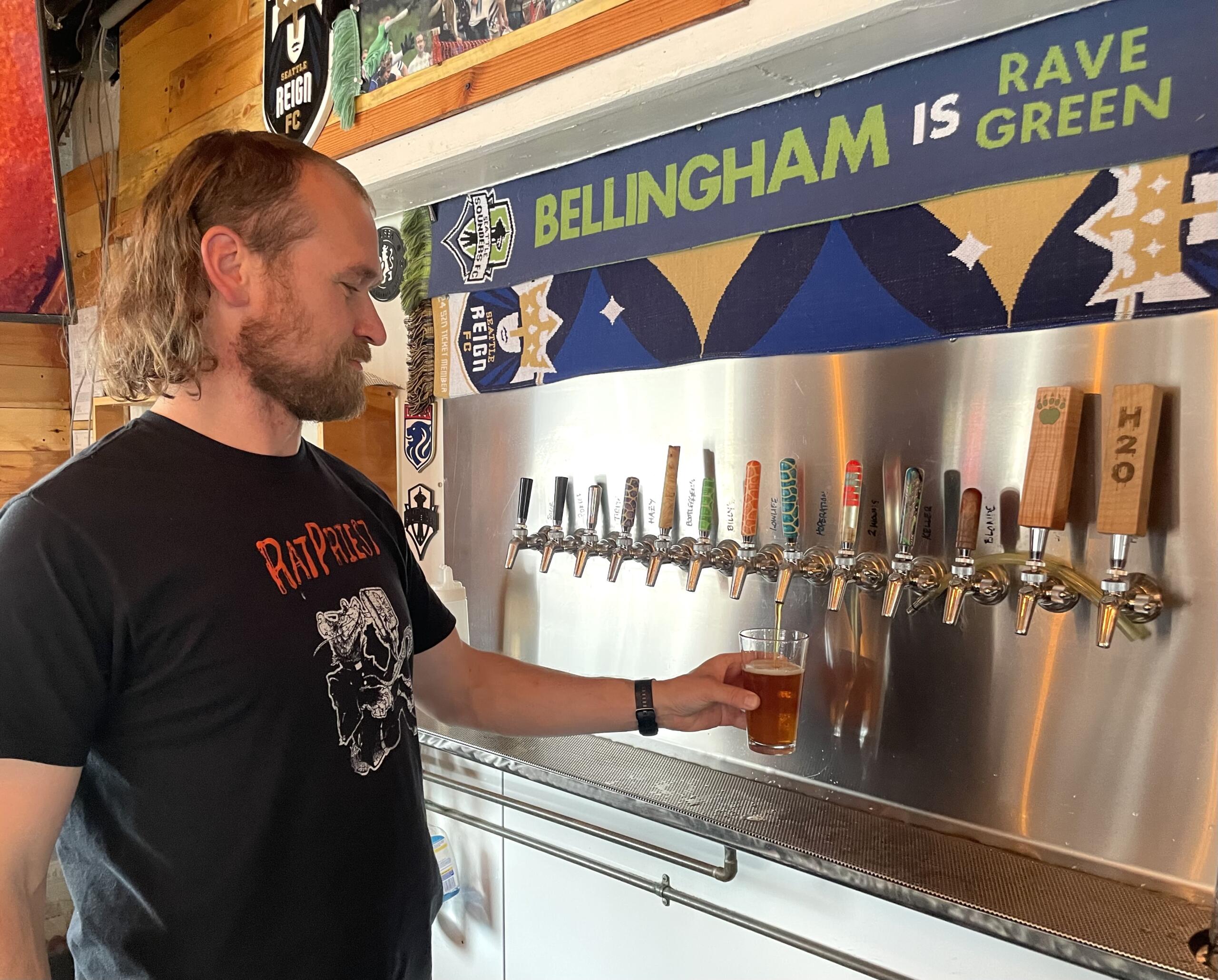 Man with long hair and beard in a black t‑shirt pouring beer into a glass at a row of taps in a bar, with a blue banner reading 'BELLINGHAM IS GREEN' in the background
