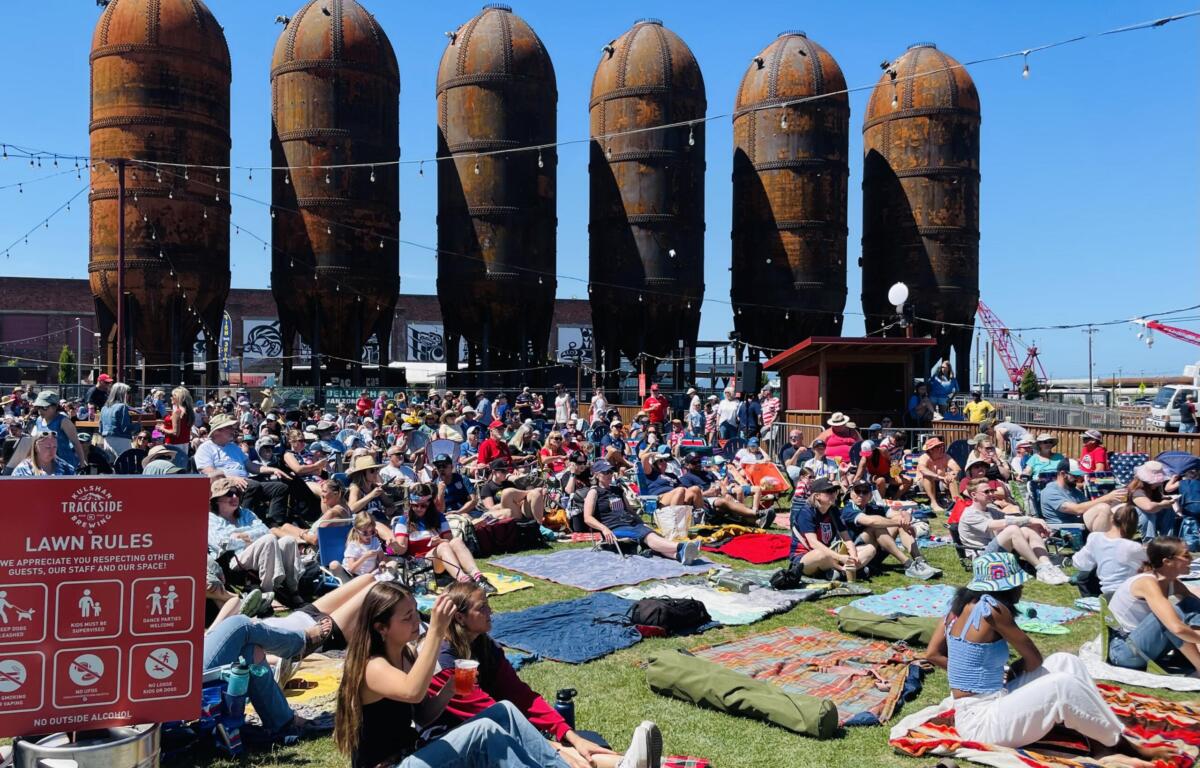 Crowd seated on blankets at an outdoor festival with rusted silo structures in the background and string lights overhead.