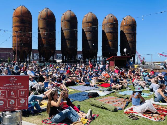 Crowd seated on blankets at an outdoor festival with rusted silo structures in the background and string lights overhead.