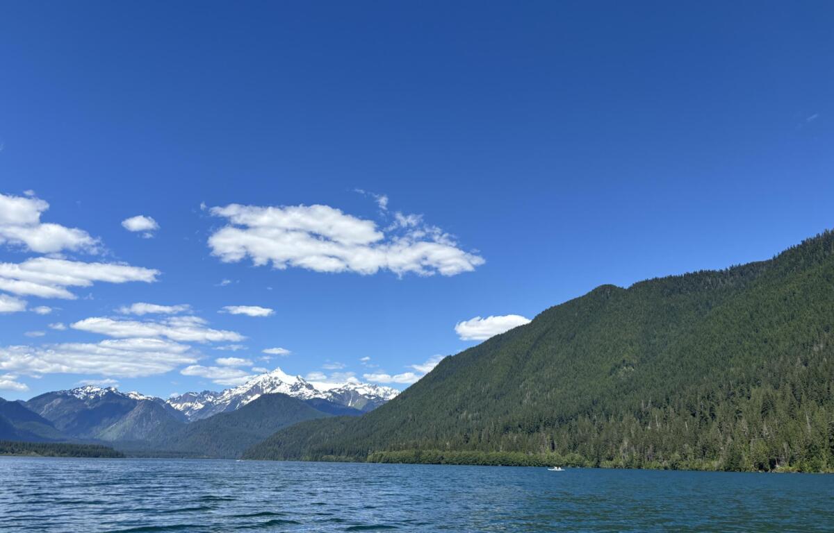 A peaceful mountain lake scene with pine-covered hills, snow-capped peaks in the distance, and a clear blue sky with a few clouds.