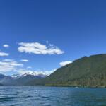 A peaceful mountain lake scene with pine-covered hills, snow-capped peaks in the distance, and a clear blue sky with a few clouds.