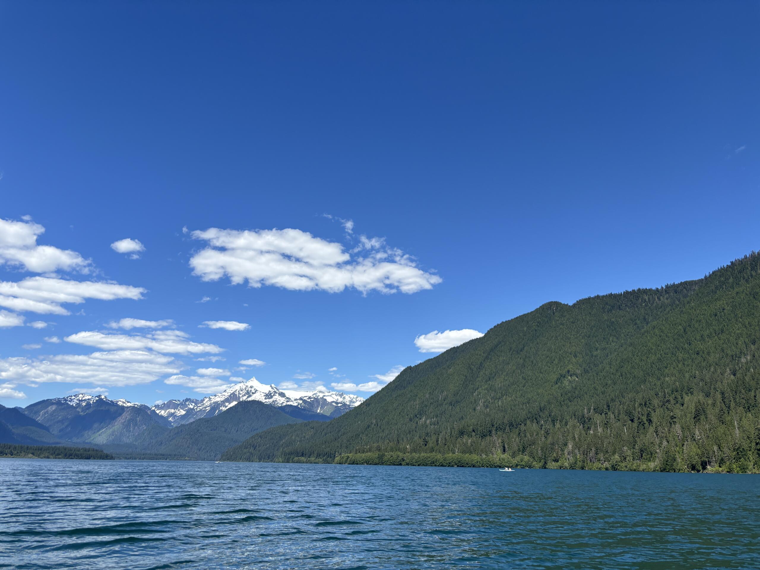 A peaceful mountain lake scene with pine-covered hills, snow-capped peaks in the distance, and a clear blue sky with a few clouds.