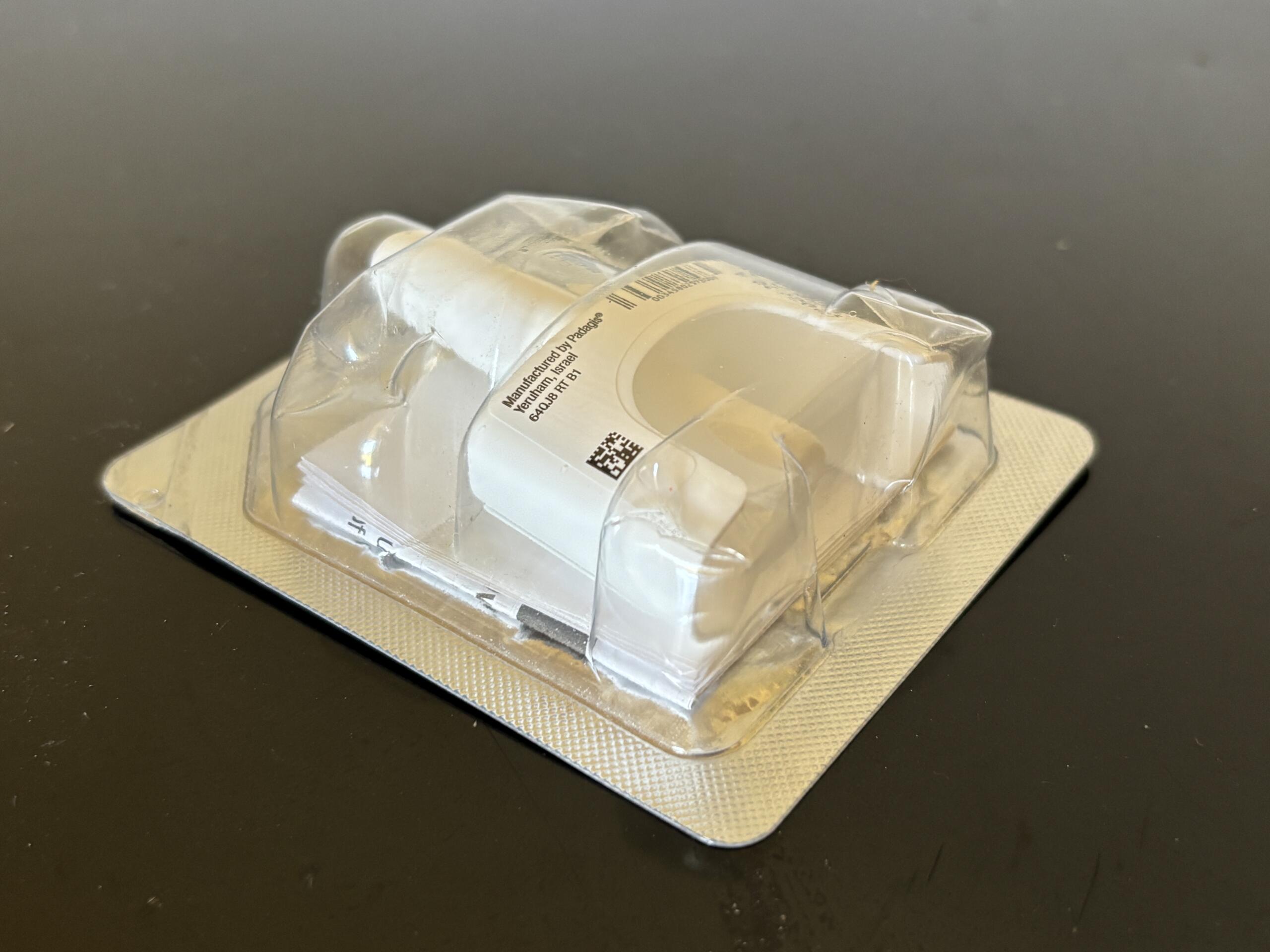 Close-up of a clear blister-pack container on a textured backing, with a partially visible label and barcode and a few white pills inside the cavitys.