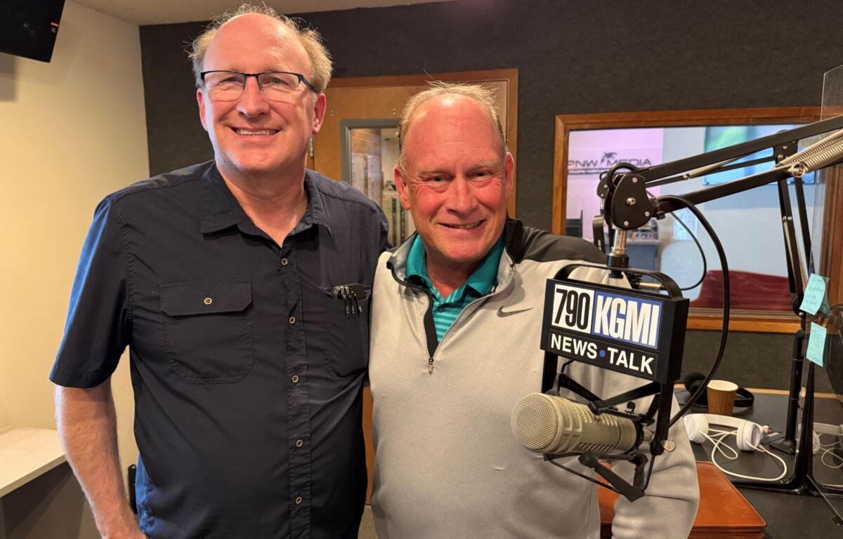 Two men pose in a radio studio with microphones and KGMI News Talk signage in view.