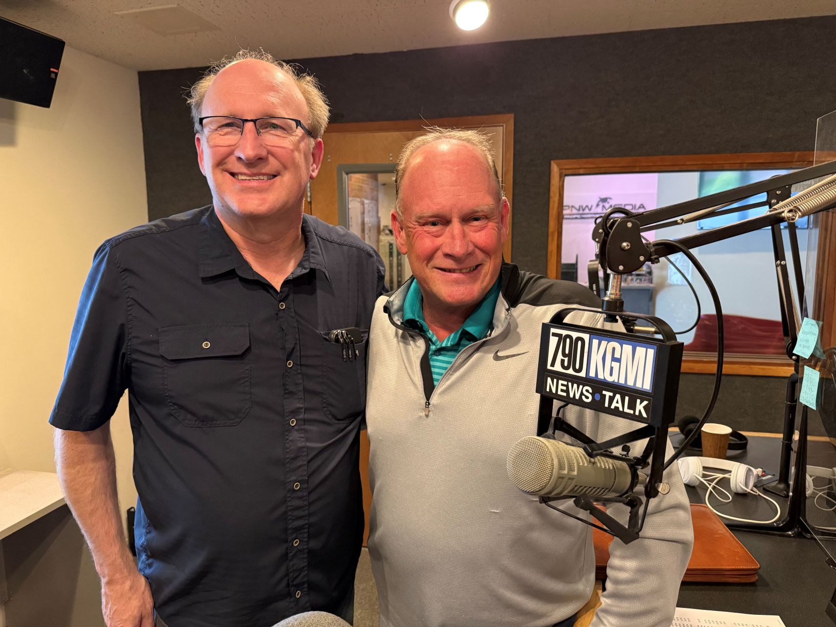 Two men pose in a radio studio with microphones and KGMI News Talk signage in view.