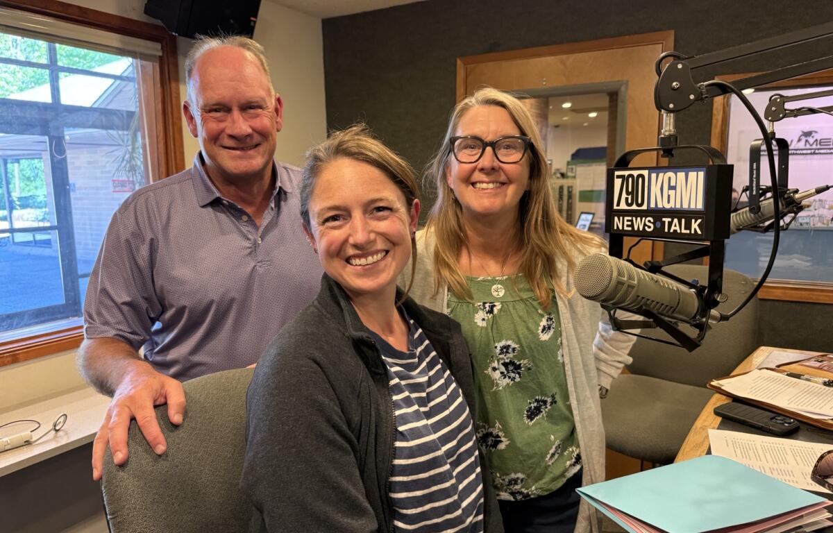 Three smiling adults posing in a radio studio with microphones and a KGMI News Talk sign in the foreground