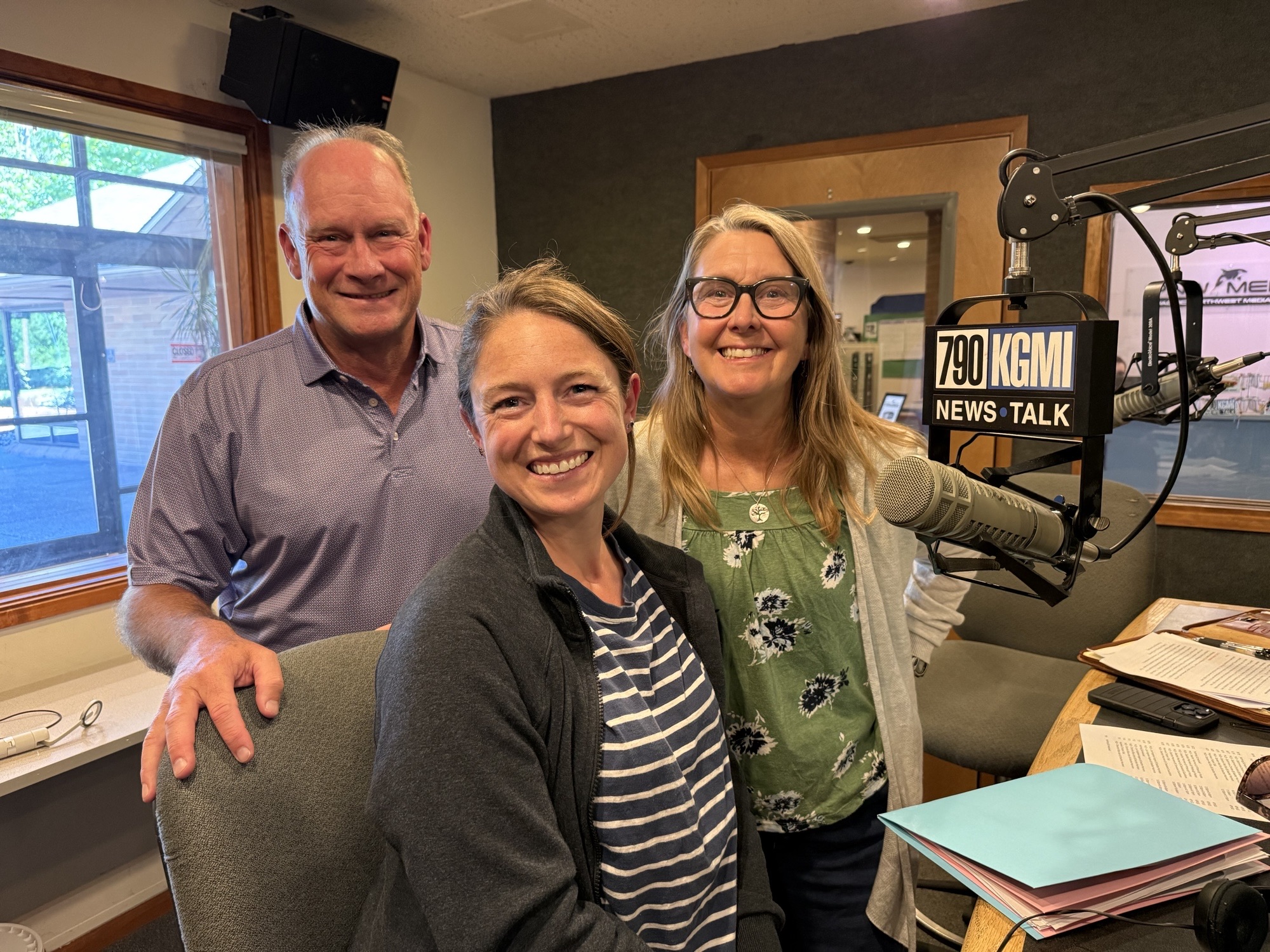 Three smiling adults posing in a radio studio with microphones and a KGMI News Talk sign in the foreground