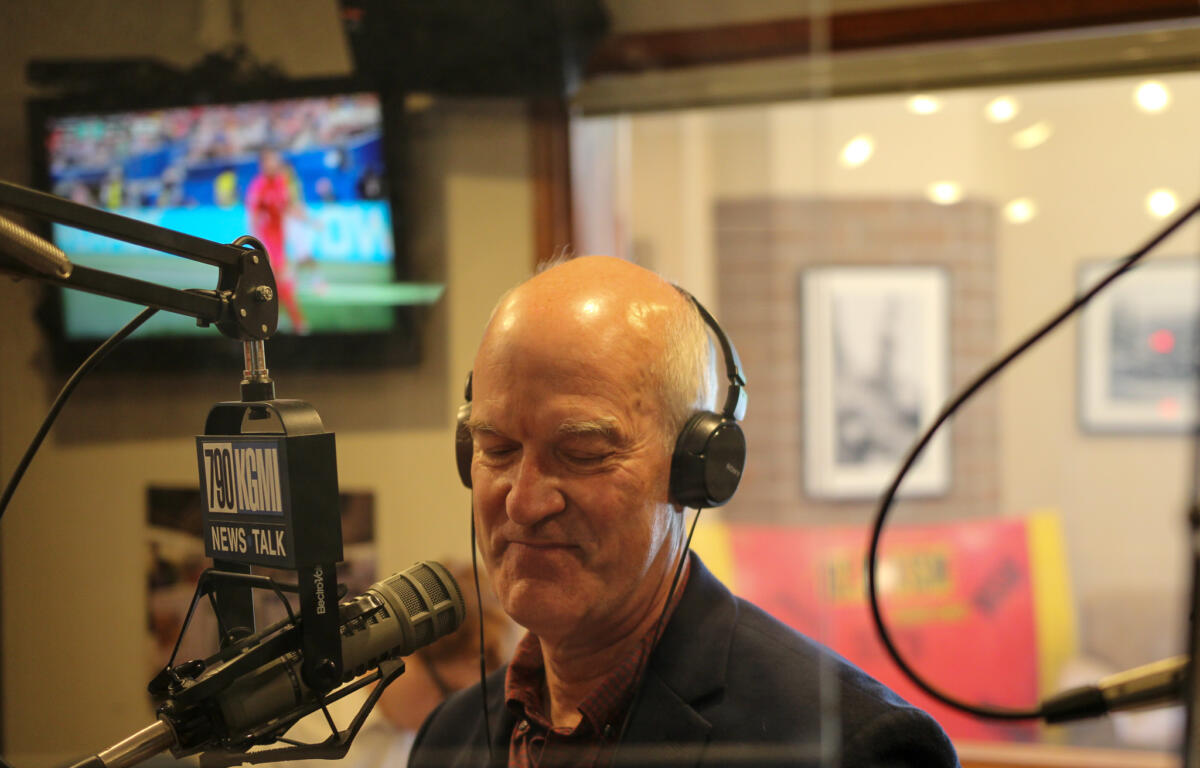 Man in a headset speaks into a professional mic in a radio studio with a 700 WLW News Talk sign visible in the foreground