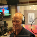 Man in a headset speaks into a professional mic in a radio studio with a 700 WLW News Talk sign visible in the foreground