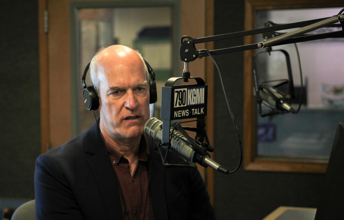 Radio host wearing headphones and speaking into a microphone in a studio with a 790 KGMN News Talk sign nearby