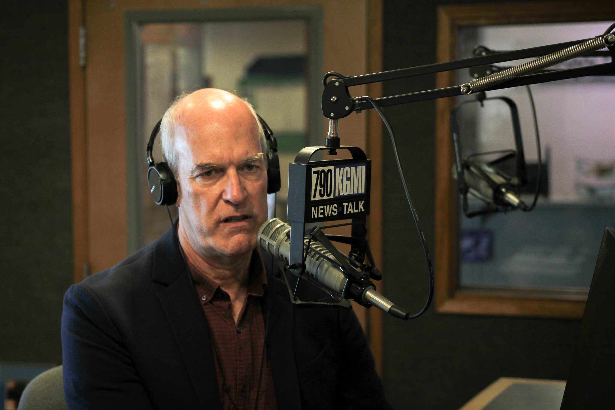 Radio host wearing headphones and speaking into a microphone in a studio with a 790 KGMN News Talk sign nearby