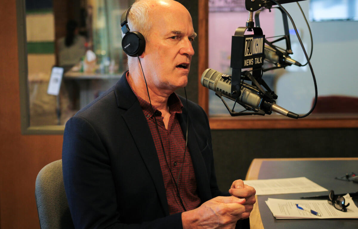 Man in a dark blazer wearing large headphones, speaking into a professional radio microphone in a studio with papers on the desk