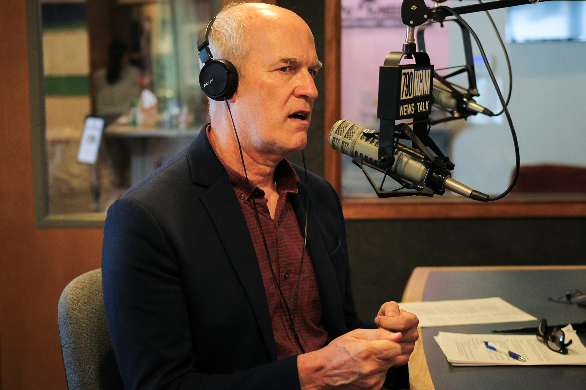 Man in a dark blazer wearing large headphones, speaking into a professional radio microphone in a studio with papers on the desk