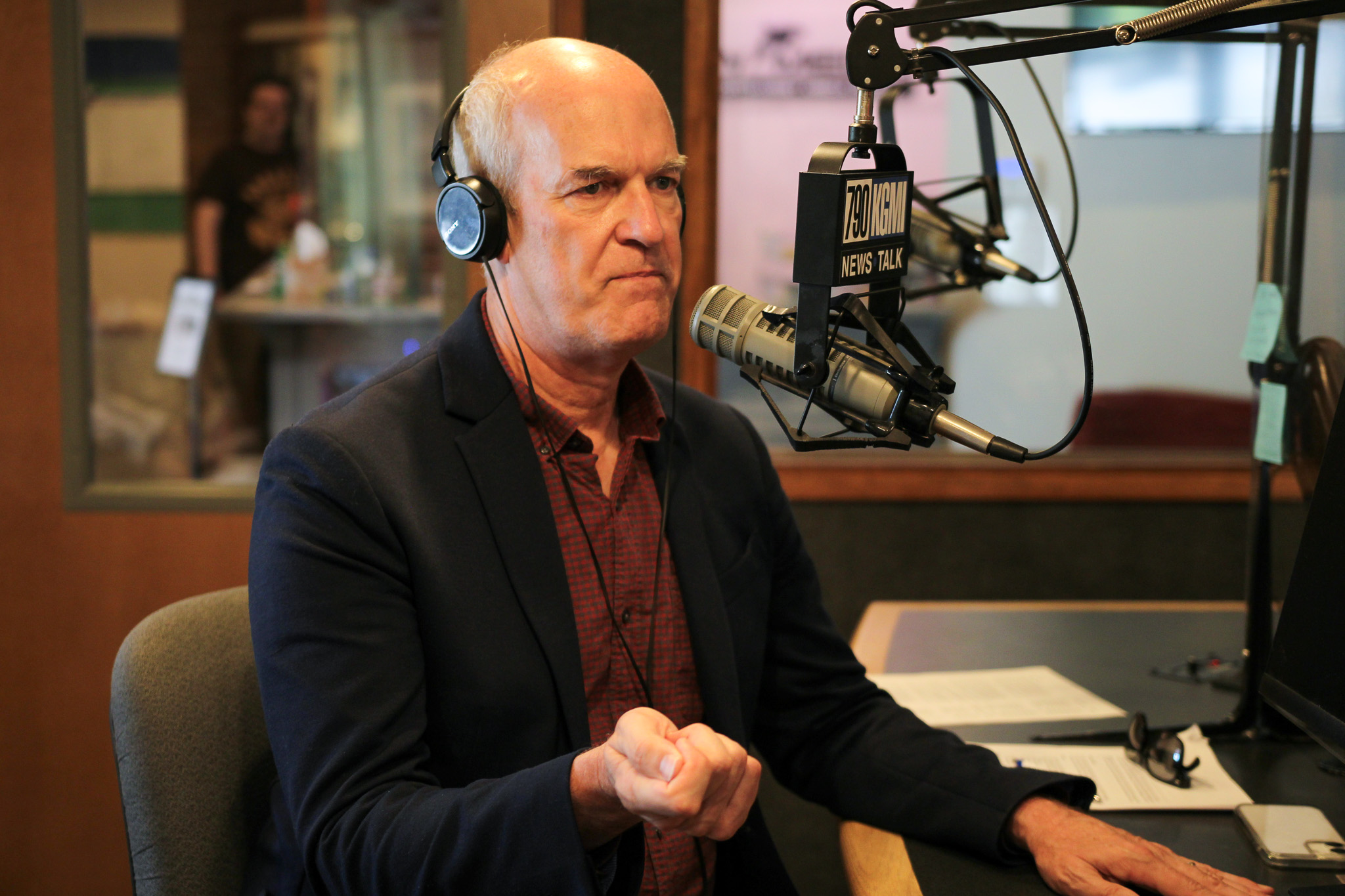 Radio host wearing headphones and a dark blazer speaks into a professional microphone in a studio, with a '790 KGM News Talk' sign on the mic front.
