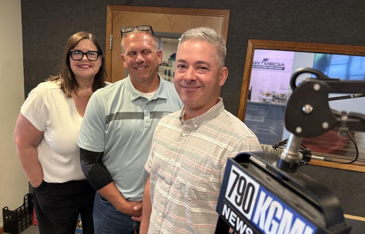 Three smiling adults pose for a photo in a radio studio, with a large microphone and “NEWS” banner in the foreground.