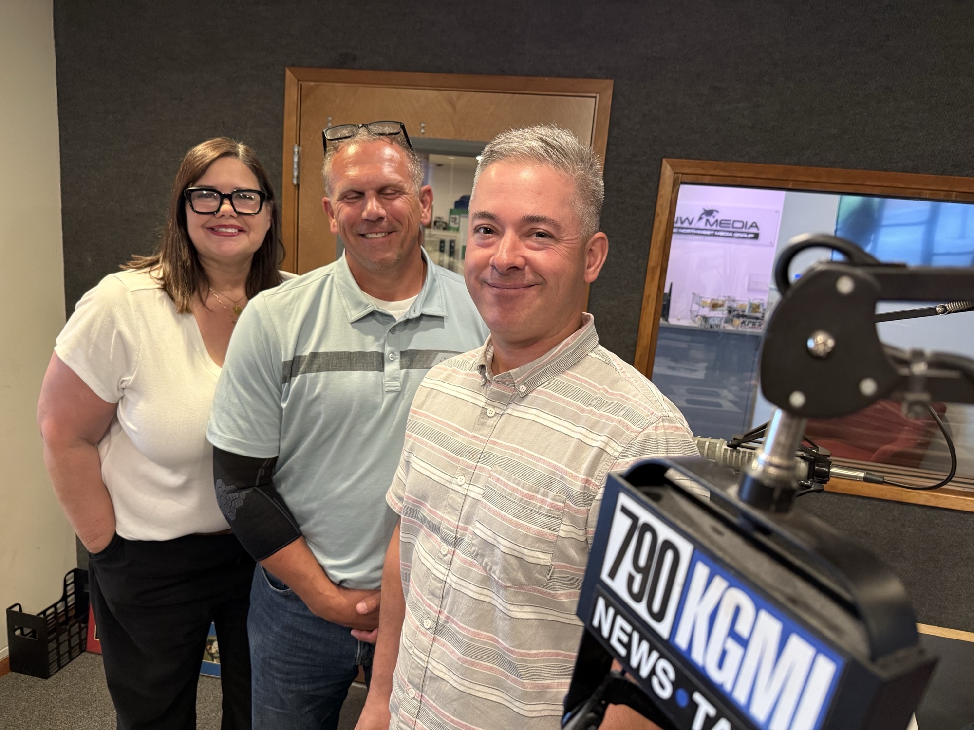 Three smiling adults pose for a photo in a radio studio, with a large microphone and “NEWS” banner in the foreground.