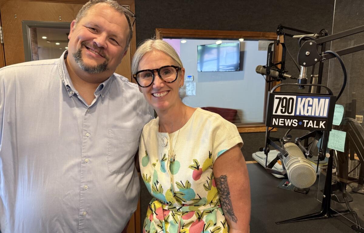 Two smiling radio hosts pose for a selfie in a studio with a large microphone and broadcast equipment visible behind them.