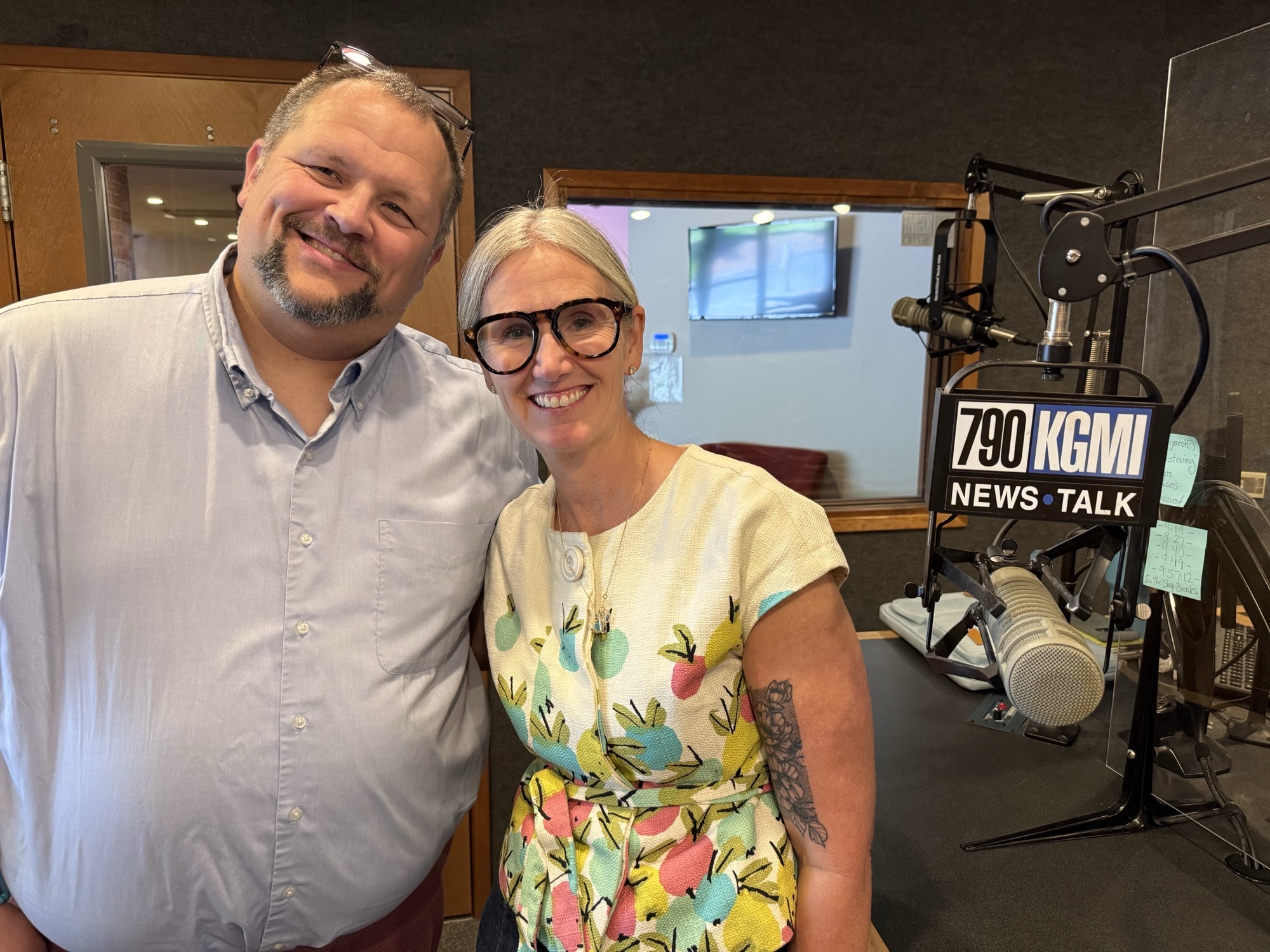 Two smiling radio hosts pose for a selfie in a studio with a large microphone and broadcast equipment visible behind them.