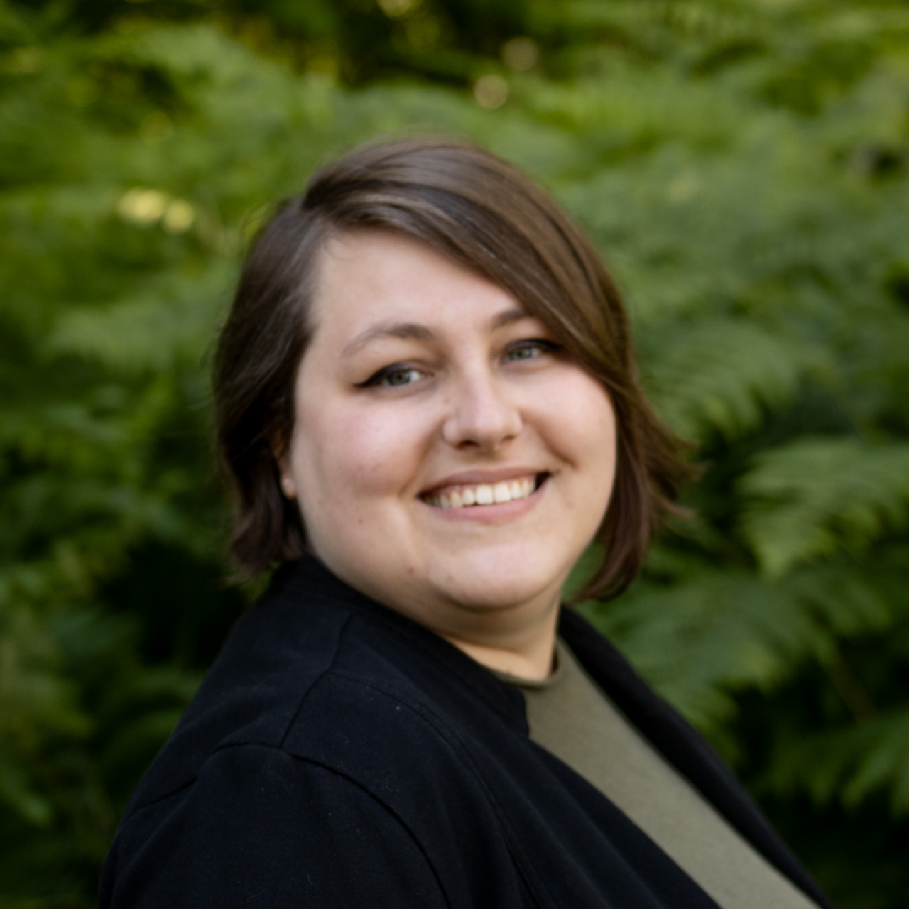 Smiling woman with short brown hair pose outdoors with green foliage in the background.