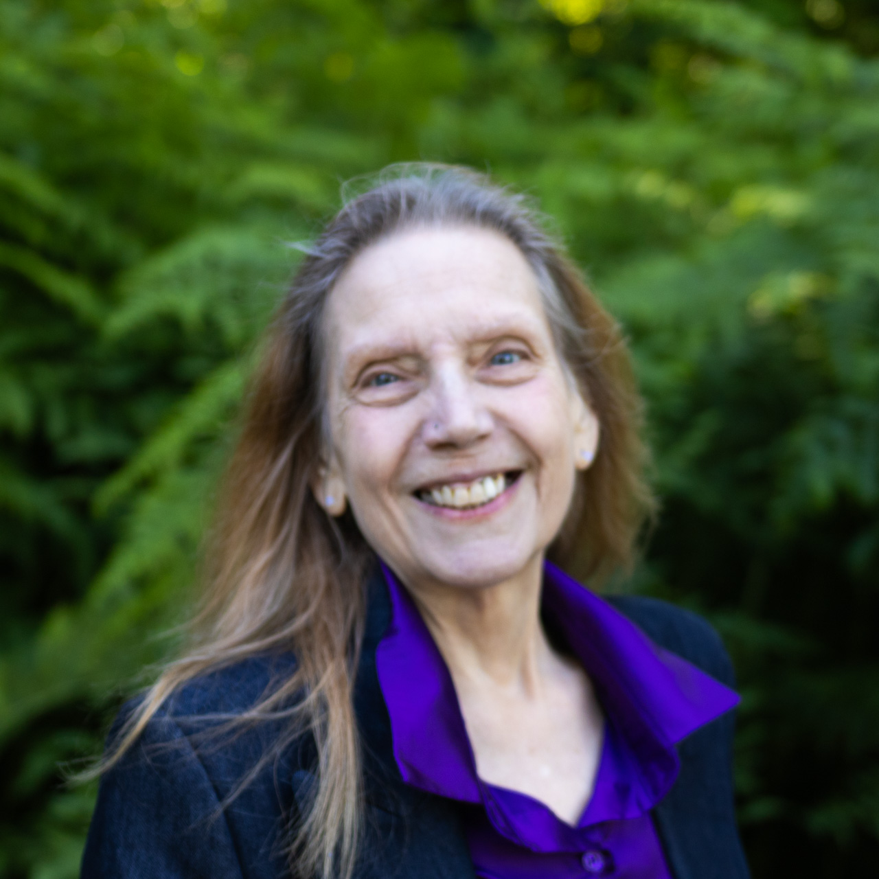 Smiling woman with long, light brown hair wearing a purple blouse and dark blazer, outdoors among green foliage.