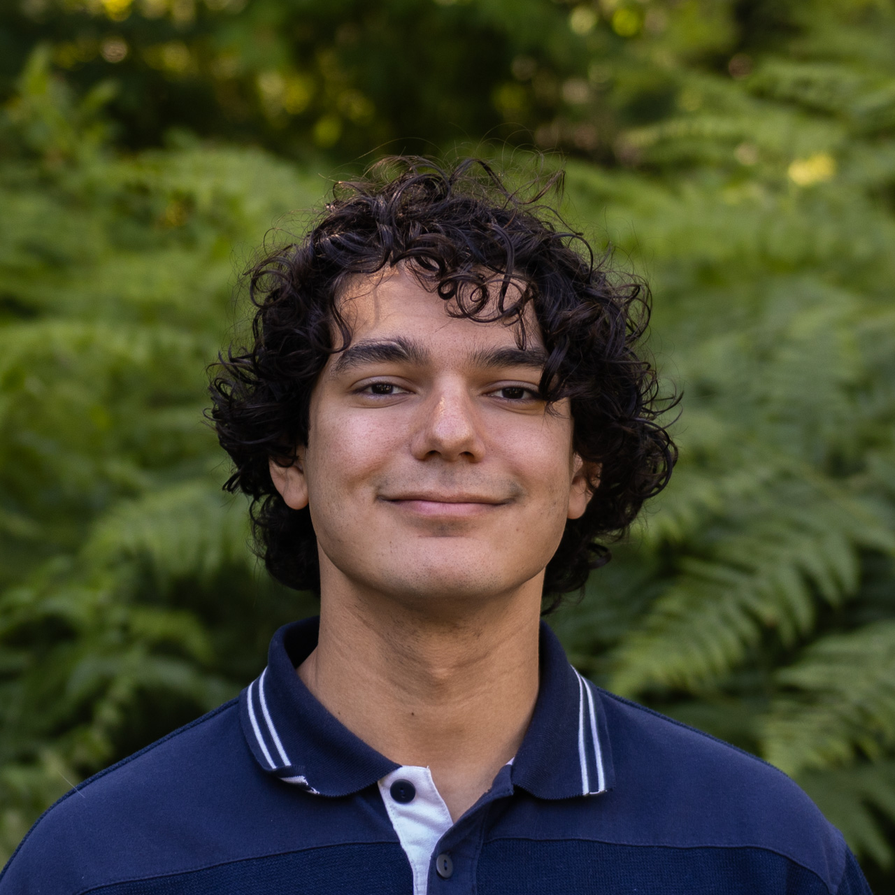 Smiling young man with curly dark hair wearing a navy polo shirt, outdoors in greenery.