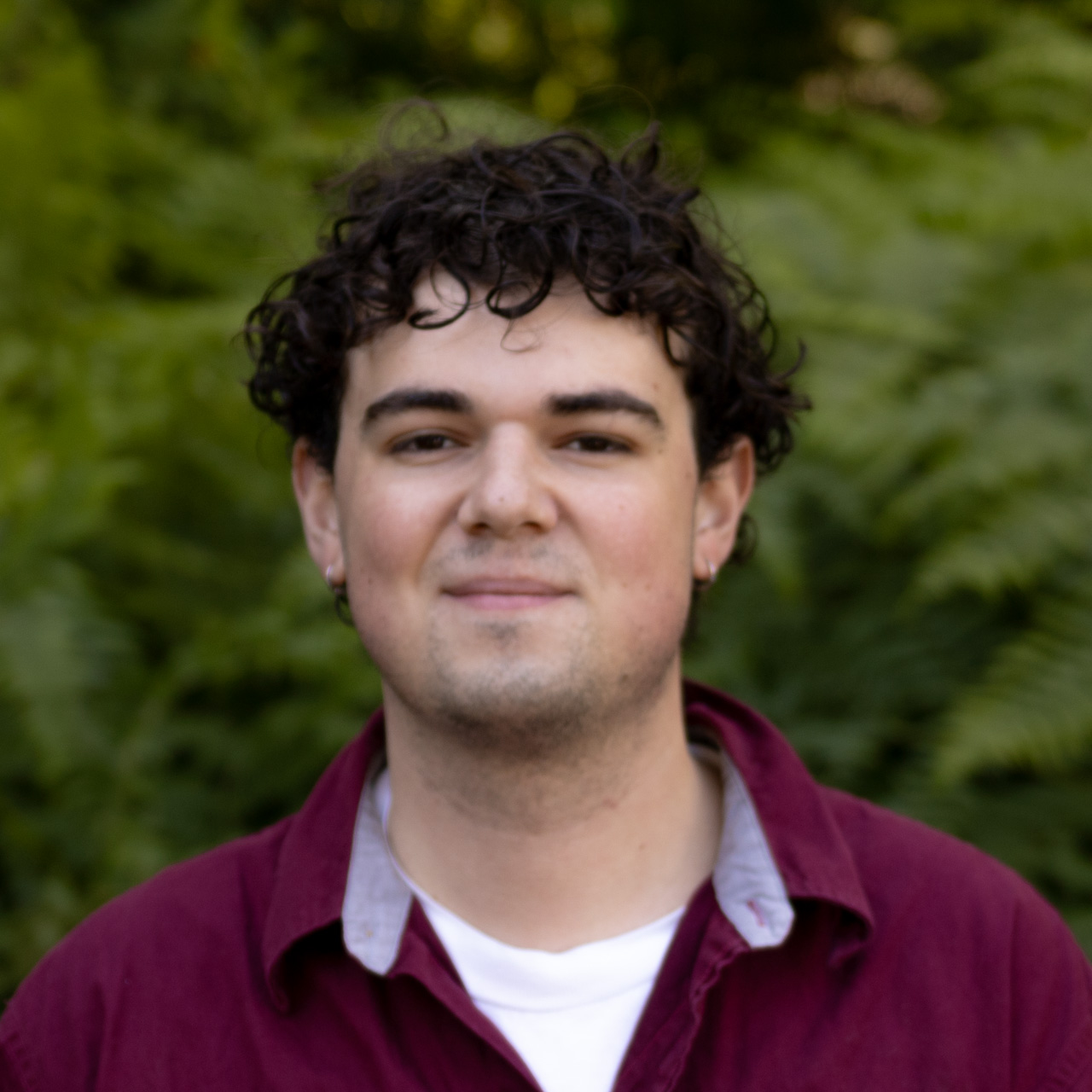 Young man with curly dark hair and a burgundy shirt smiles gently with a green foliage background behind him.