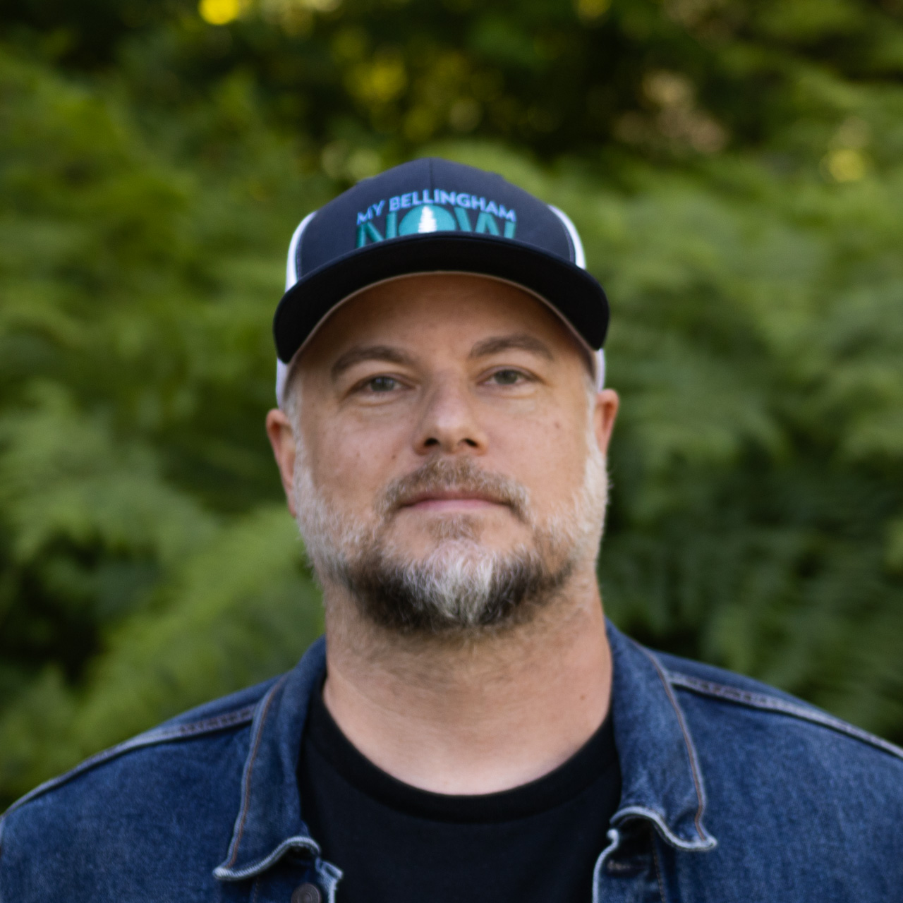 Portrait of a man with a gray beard wearing a dark cap that reads 'My Bellingham Now' and a denim jacket outdoors.