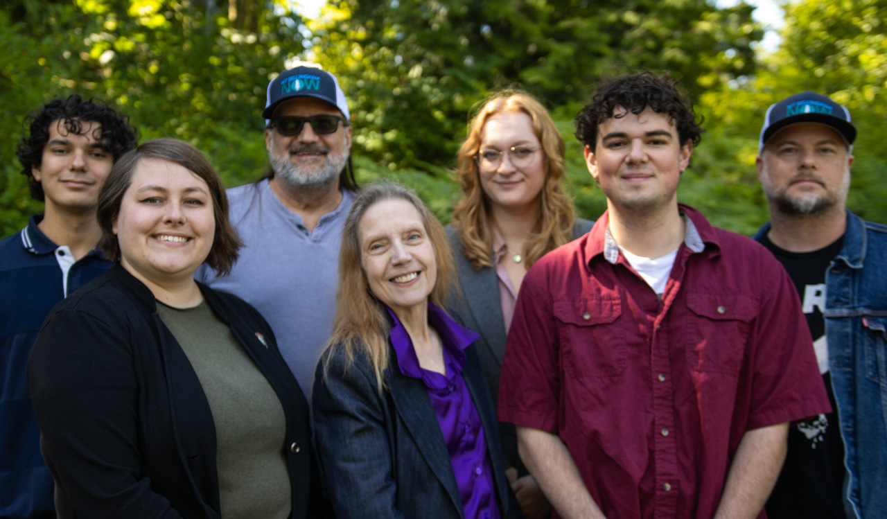 Group of eight adults smiling outdoors, posing for a team photo in a sunny park.