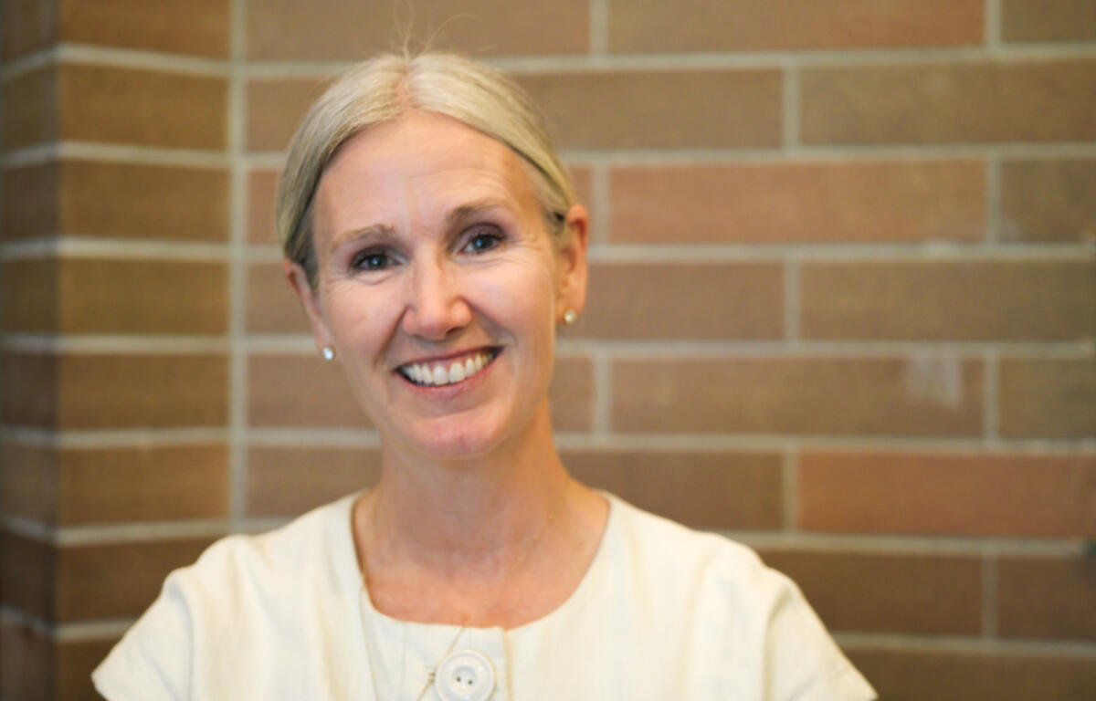 Smiling older woman with short gray hair in a light blouse standing against a brick wall, looking at the camera.