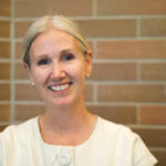 Smiling older woman with short gray hair in a light blouse standing against a brick wall, looking at the camera.