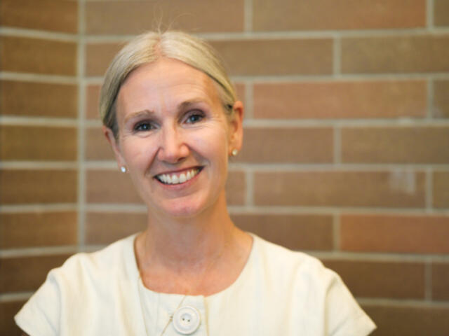 Smiling older woman with short gray hair in a light blouse standing against a brick wall, looking at the camera.
