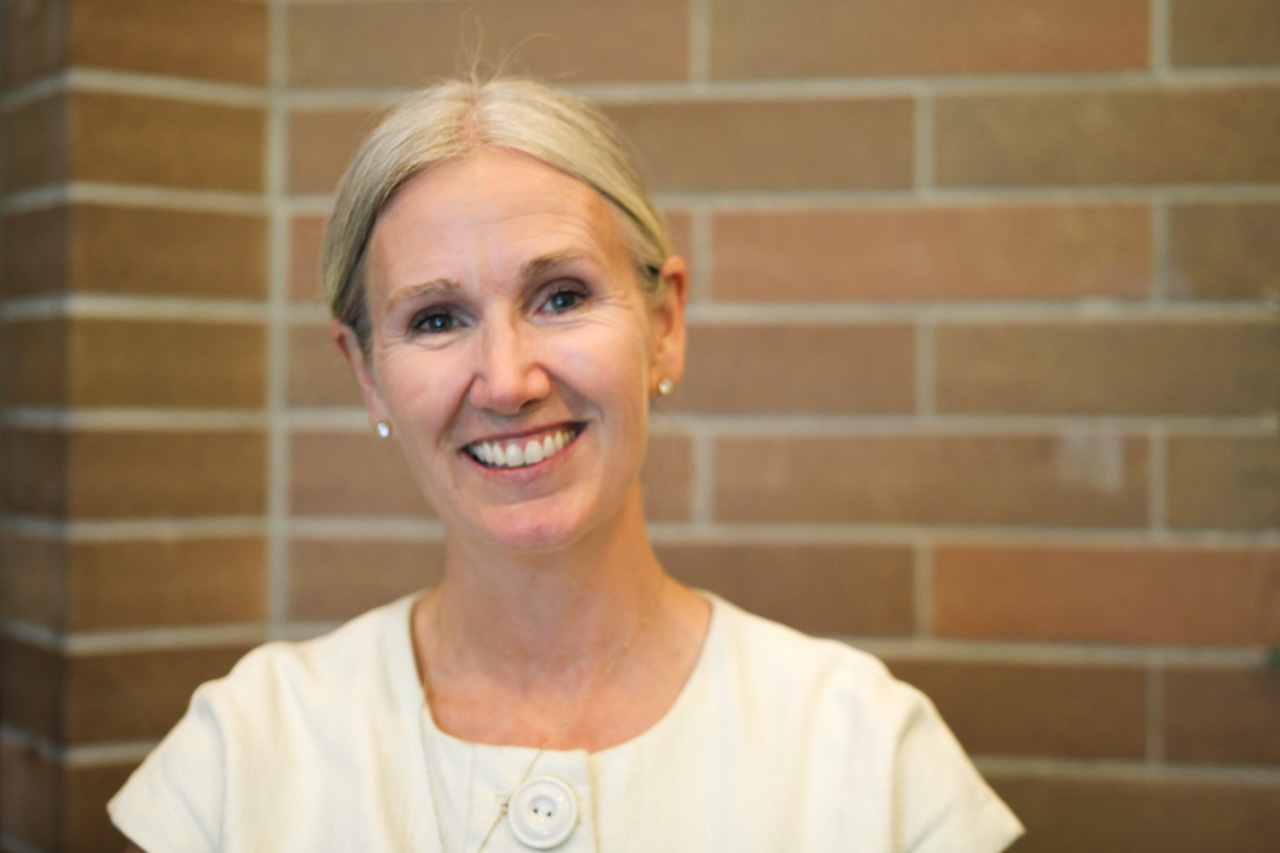 Smiling older woman with short gray hair in a light blouse standing against a brick wall, looking at the camera.