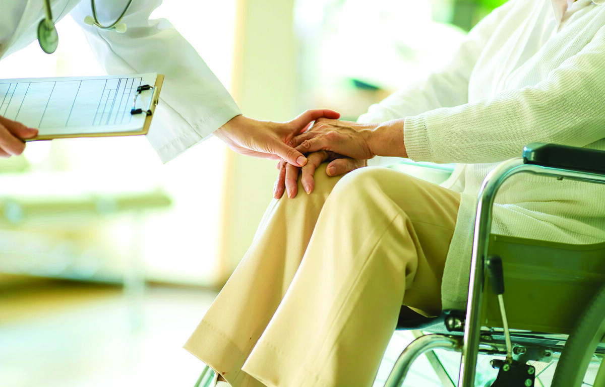 A clinician holds an elderly patient’s hand in a wheelchair, conveying care and support. A clipboard is visible nearby for notes.