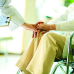 A clinician holds an elderly patient’s hand in a wheelchair, conveying care and support. A clipboard is visible nearby for notes.