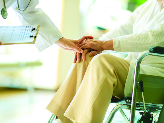 A clinician holds an elderly patient’s hand in a wheelchair, conveying care and support. A clipboard is visible nearby for notes.